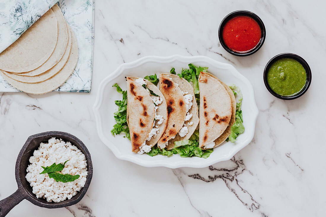 Tortillas with fresh quark cheese placed on plate with lettuce leaves, bowls of avocado and tomato sauces