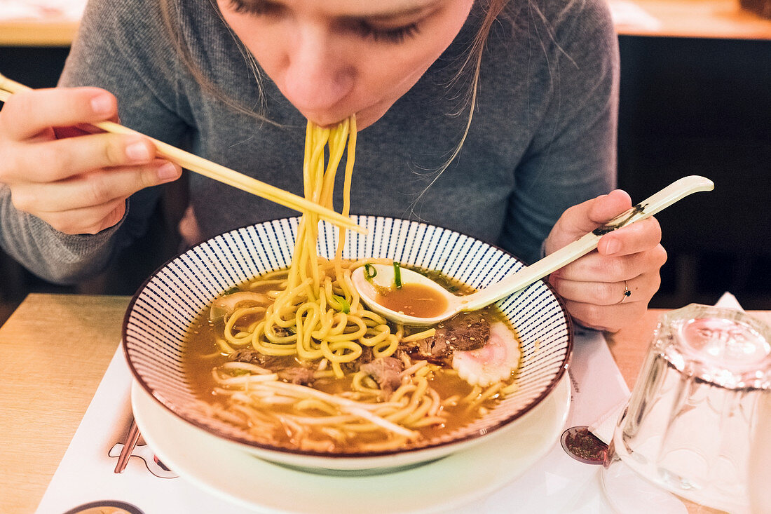 Young female using chopsticks and spoon to eat tasty ramen while sitting at table in Japanese restaurant