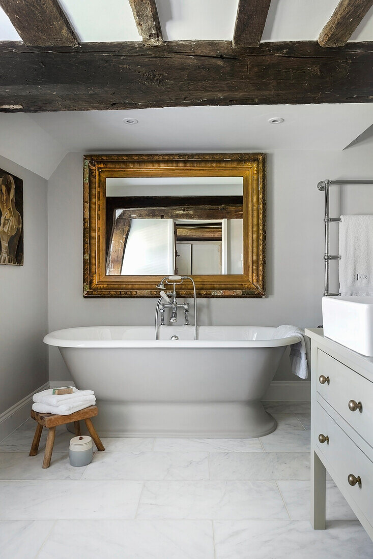 Freestanding bathtub and gold framed mirror in light grey bathroom with rustic wooden beams