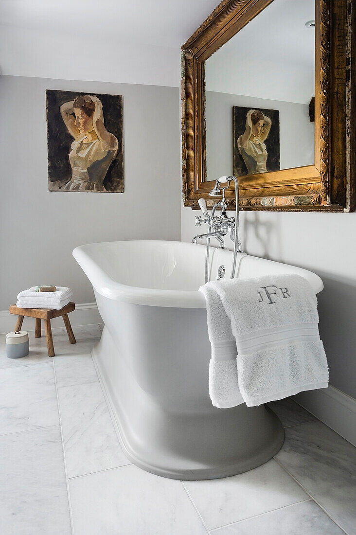 Free standing bathtub, gold framed mirror and painting above in light grey bathroom