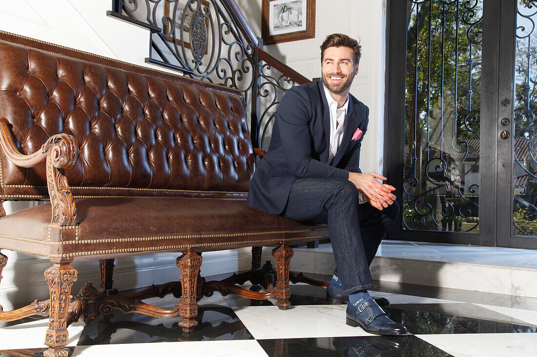 Young man with a beard in a jacket and jeans is sitting on a leather sofa