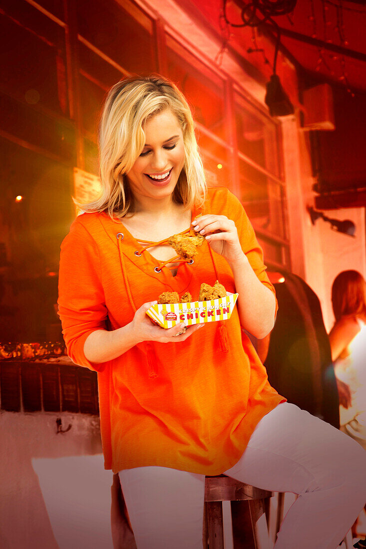 Junge blonde Frau in orangefarbener Bluse isst Fast Food