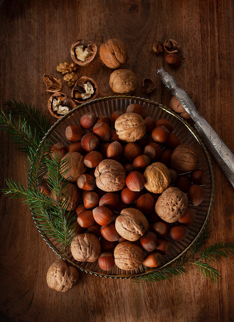 Hazelnuts and walnuts with sprigs of pine needles in a silver bowl beside a nutcracker