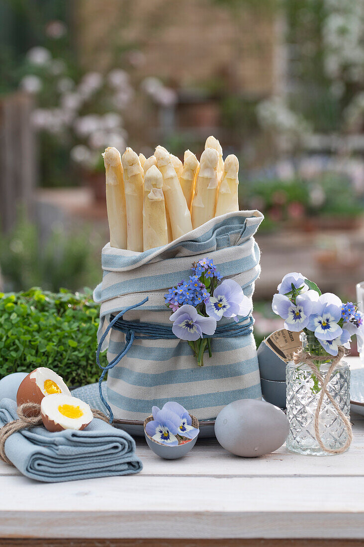 Table with asparagus tips wrapped in cloth, garden pansies (Viola wittrockiana) and forget-me-nots in vase, Easter eggs as decoration
