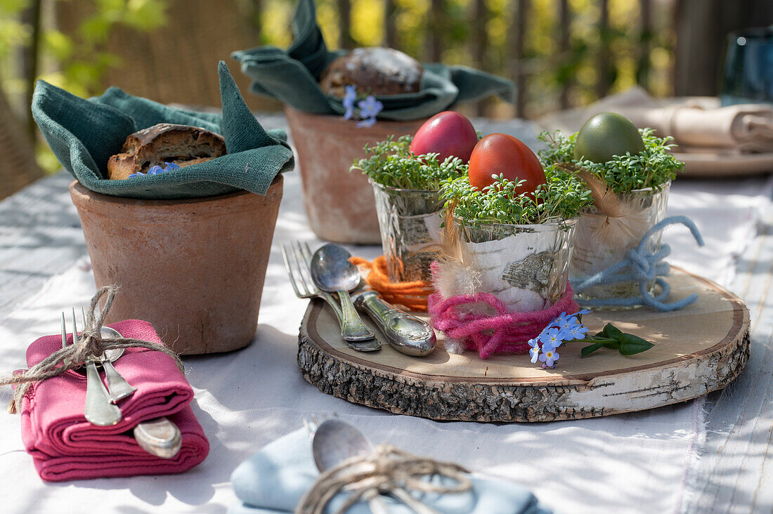 Ostereier mit Kresse in Gläsern auf Holzscheibe, Brotscheiben in Tontöpfen und Besteck am Tisch, Osterdeko