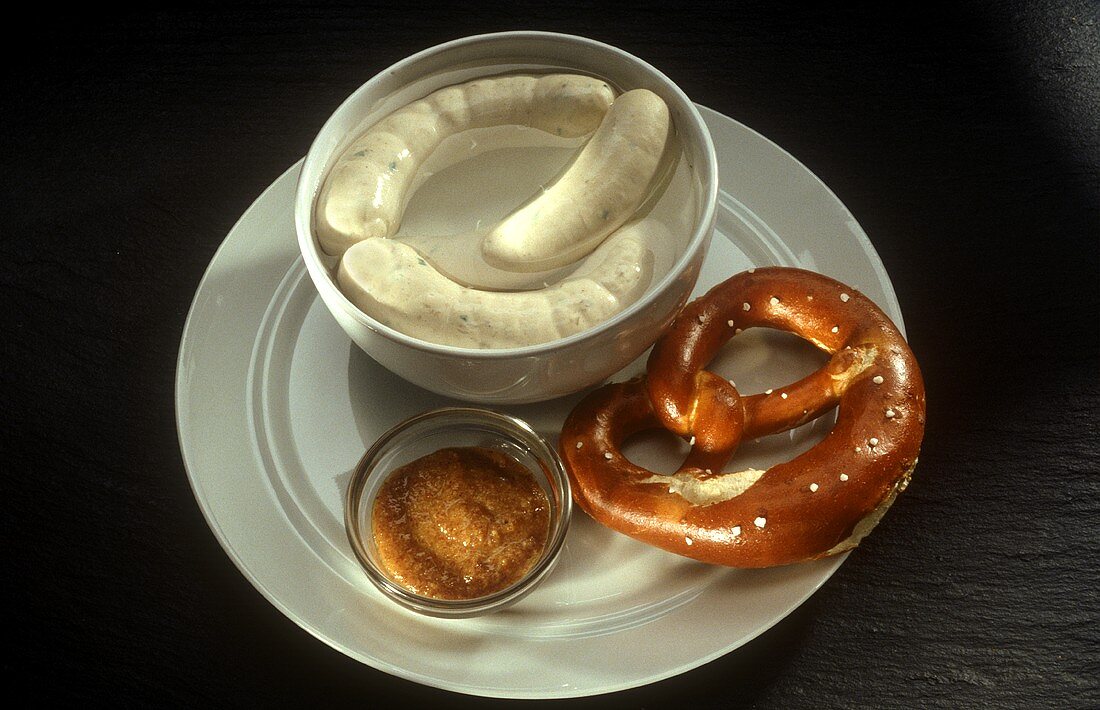 White sausages in dish, mild mustard & pretzel on plate