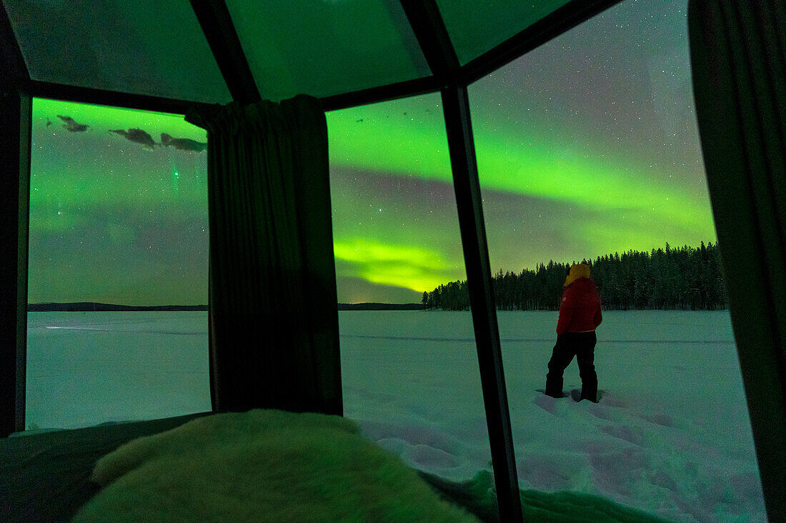 Person standing in the snow looking at Aurora Borealis (Northern Lights), view through glass windows of igloo, Jokkmokk, Lapland, Sweden, Scandinavia, Europe