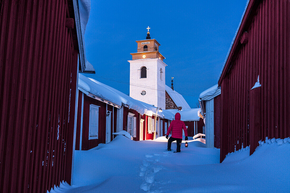 Person admiring the bell tower standing in deep snow among red cottages at dusk, Gammelstad Church Town, UNESCO World Heritage Site, Lulea, Sweden, Scandinavia, Europe