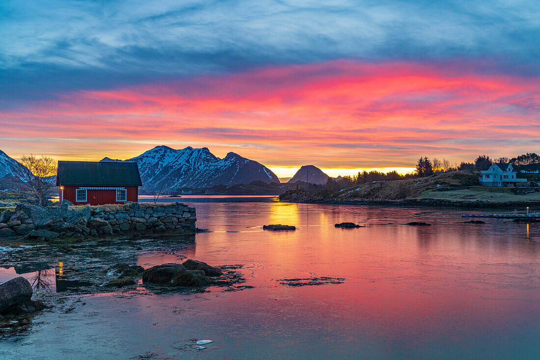 Feuriger Himmel bei Sonnenaufgang über einem roten Rorbu mit Blick auf das gefrorene Meer, Ballstad, Vestvagoy, Lofoten Inseln, Norwegen, Skandinavien, Europa