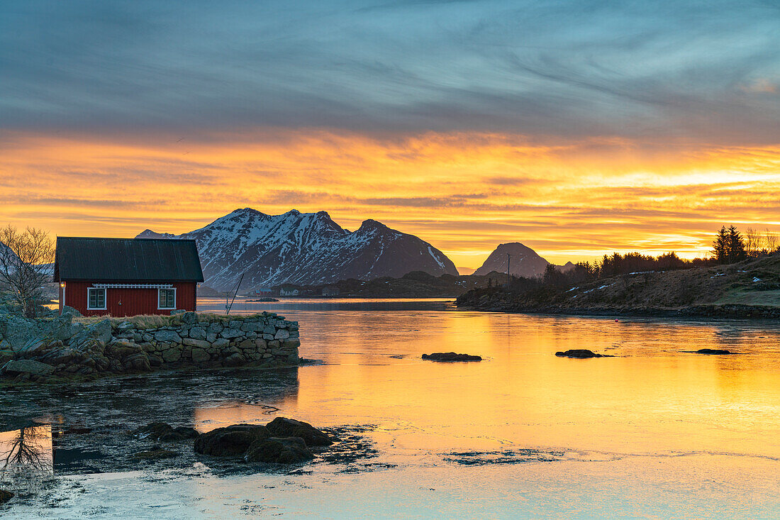 Snowcapped mountains and fisherman's red cabin under the burning sky at dawn, Ballstad, Vestvagoy, Lofoten Islands, Norway, Scandinavia, Europe