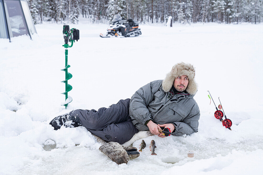 Mann liegt auf dem Eis und angelt aus einem Loch, Lappland, Schweden, Skandinavien, Europa