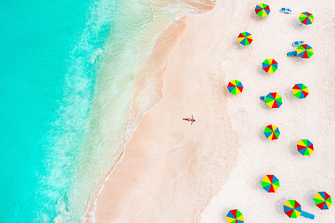 Luftaufnahme einer Frau im Bikini beim Sonnenbaden an einem tropischen Strand neben bunten Sonnenschirmen, Antigua, Westindische Inseln, Karibik, Mittelamerika