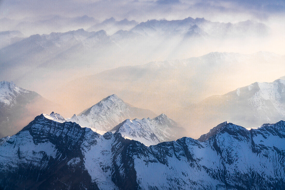Sonnenstrahlen durch Nebel über den schneebedeckten Gipfeln der Lepontiner und Tessiner Alpen bei Sonnenuntergang, Blick aus dem Flugzeug, Schweiz, Europa
