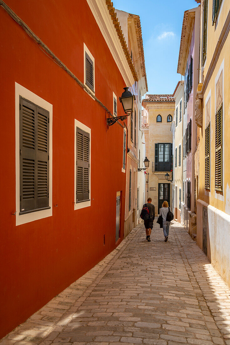 Blick auf ein Paar, das eine pastellfarbene Straße im historischen Zentrum entlanggeht, Ciutadella, Menorca, Balearen, Spanien, Mittelmeer, Europa