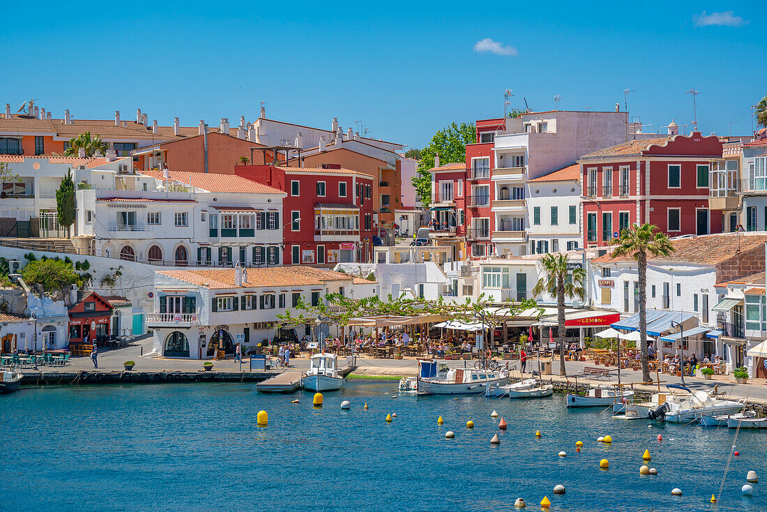 Blick auf bunte Cafés, Restaurants und Boote im Hafen vor blauem Himmel, Cales Fonts, Menorca, Balearen, Spanien, Mittelmeer, Europa
