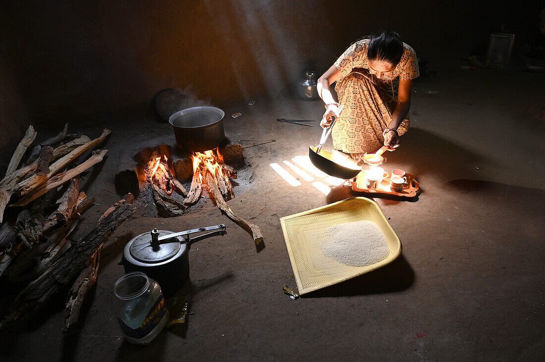 Sonnenlicht fällt in eine traditionelle Küche mit Lehmwänden und Holzfeuer, Frau macht Chai, Reissieb aus Plastik, Kadana, Gujarat, Indien, Asien