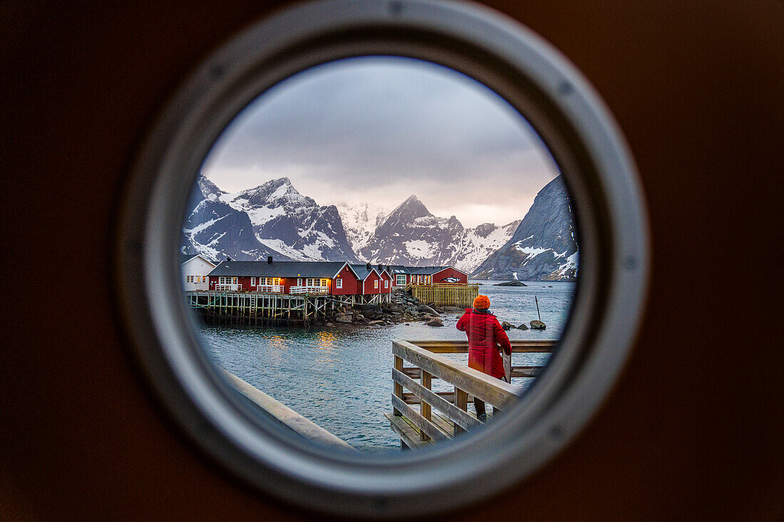 Rückansicht einer Person, die durch ein Bullauge auf traditionelle Rorbu-Hütten blickt, Reine-Bucht, Lofoten-Inseln, Norwegen, Skandinavien, Europa
