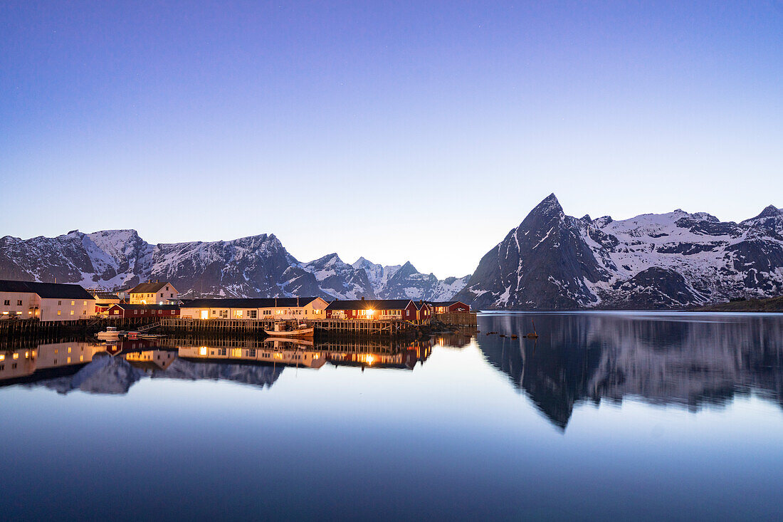 Fischerdorf, das sich in der Abenddämmerung im Meer spiegelt, mit schneebedeckten Bergen im Hintergrund, Hamnoy, Reine-Bucht, Lofoten-Inseln, Norwegen, Skandinavien, Europa