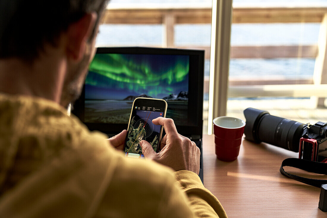Ein Mann schaut sich Karten auf seinem Smartphone an, während er mit seinem Laptop arbeitet, Lofoten Inseln, Norwegen, Skandinavien, Europa