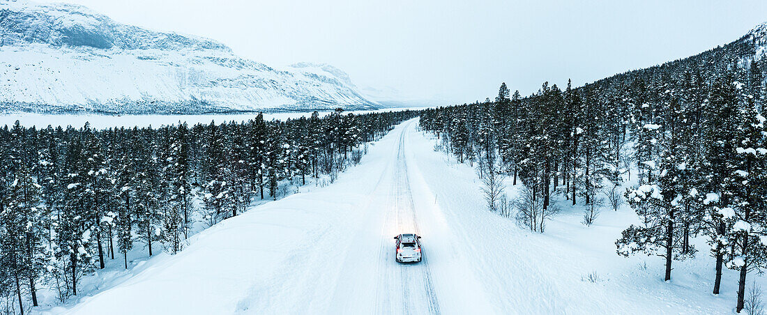 Blick von oben auf ein Auto, das auf einer vereisten Straße durch einen schneebedeckten Wald fährt, Stora Sjofallet, Landkreis Norrbotten, Lappland, Schweden, Skandinavien, Europa