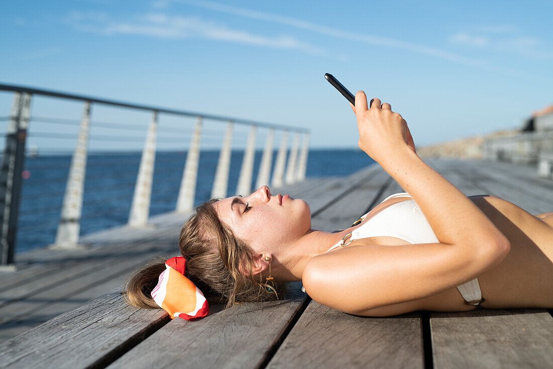 Junge Frau liegt auf einem Steg und hält ein Handy in der Hand