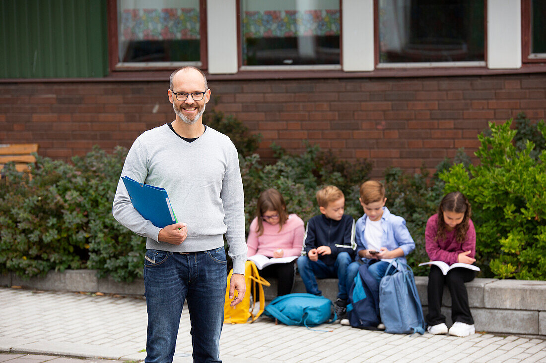 Lächelnde Lehrerin vor der Schule, Schulkinder im Hintergrund