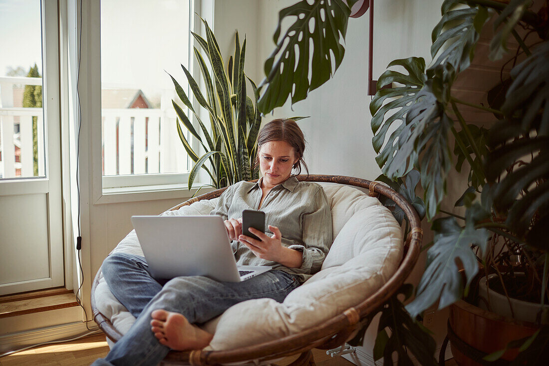 Frau im Papasan-Stuhl sitzend, Laptop und Telefon benutzend