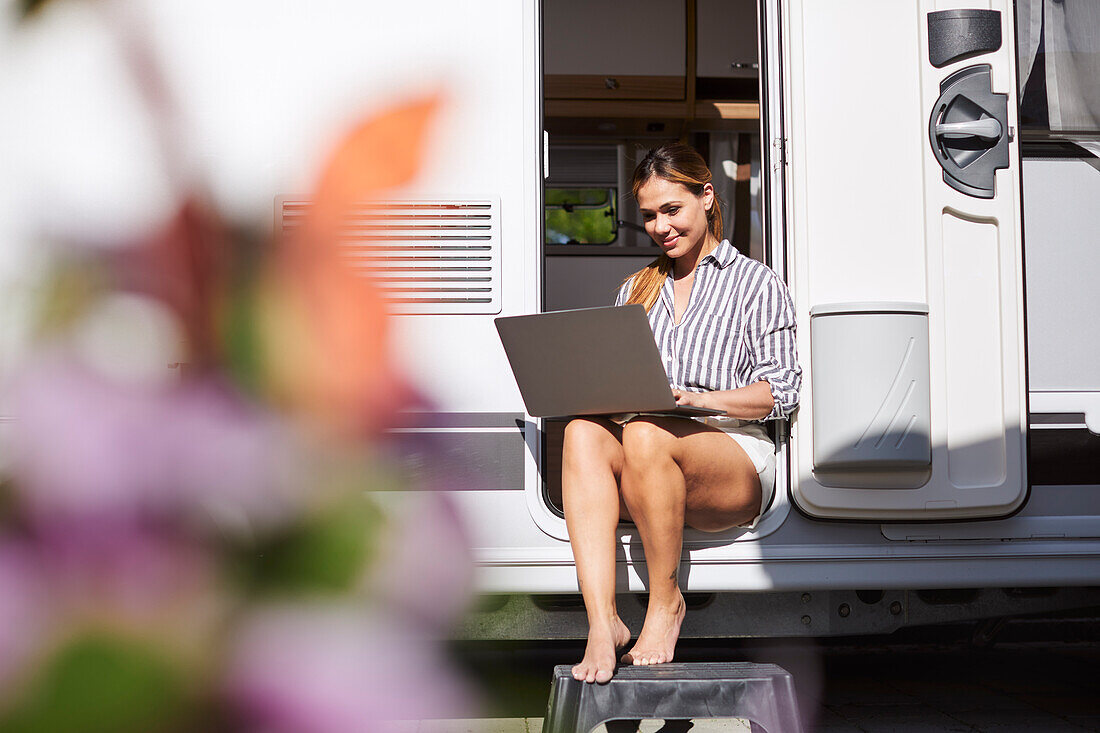 Frau sitzt an der Tür eines Wohnwagens und benutzt einen Laptop