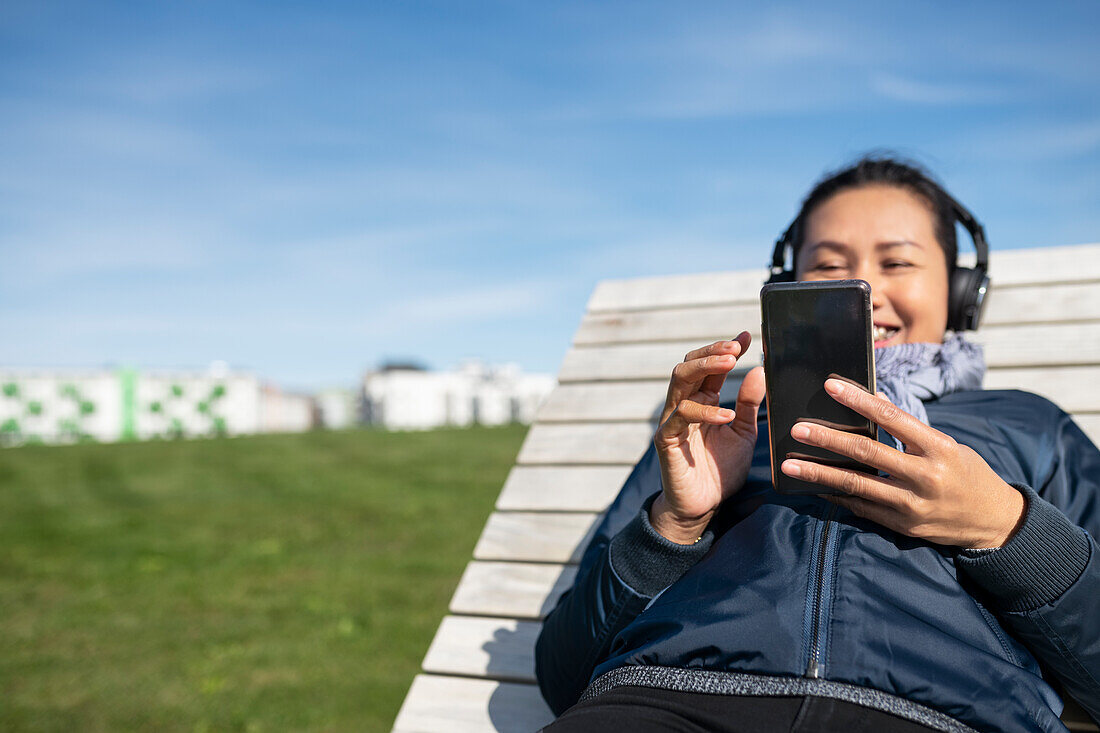 Junge Frau mit Smartphone entspannt sich auf einem hölzernen Sonnenstuhl