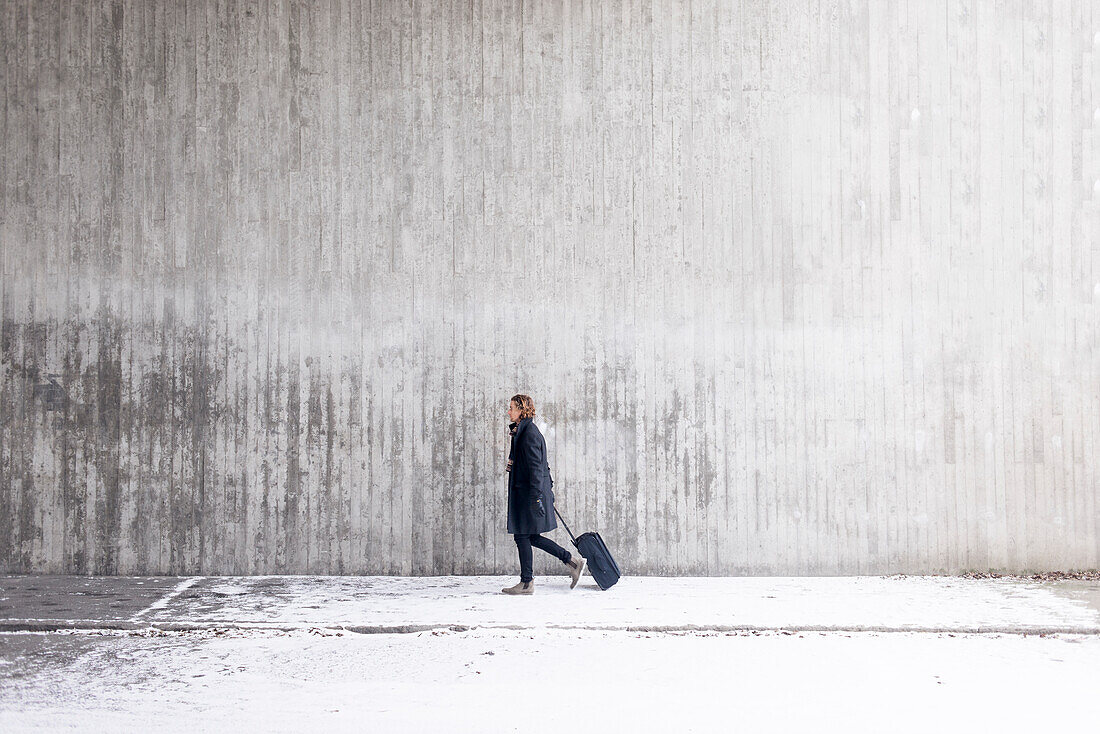 Junger Mann mit Koffer geht im Winter an einer Mauer entlang