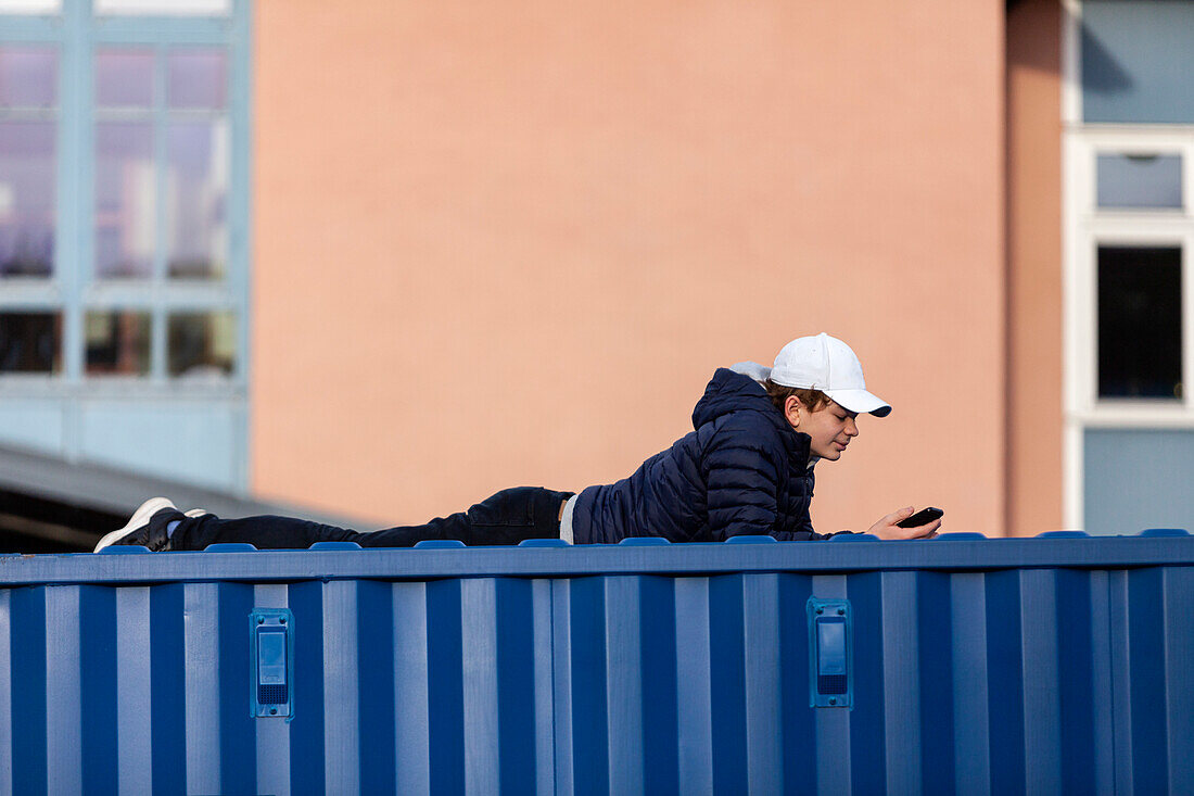 Teenage boy lying on shipping container and using cell phone