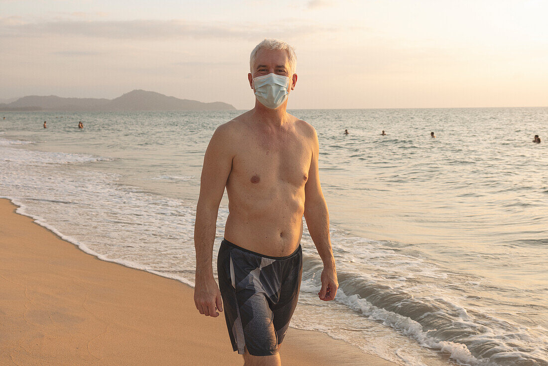 Älterer Mann, der mit einer Gesichtsmaske am Strand spazieren geht