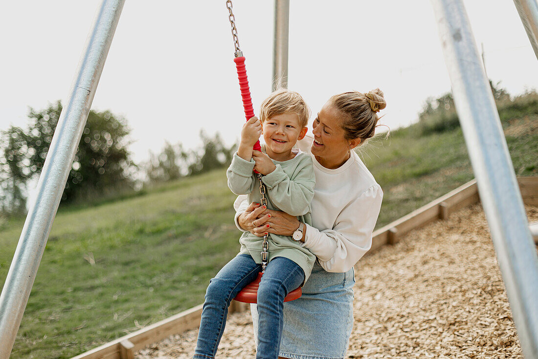 Glückliche Mutter schaukelt ihren Sohn auf dem Spielplatz