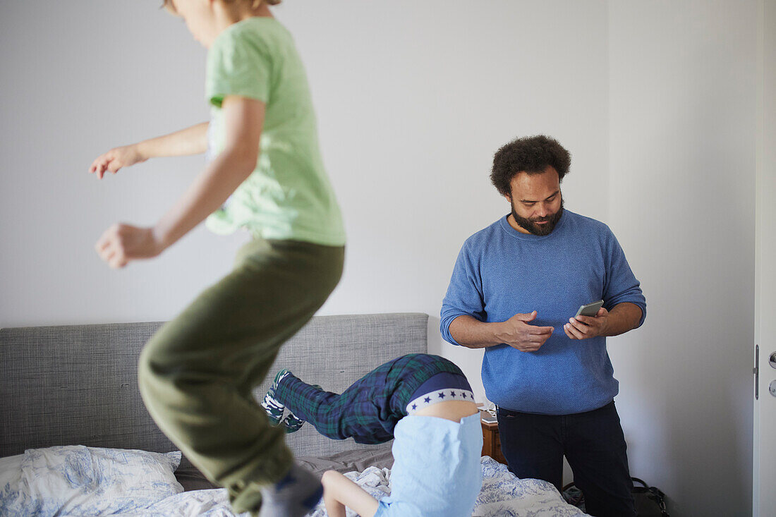 Father using phone while sons jumping on bed