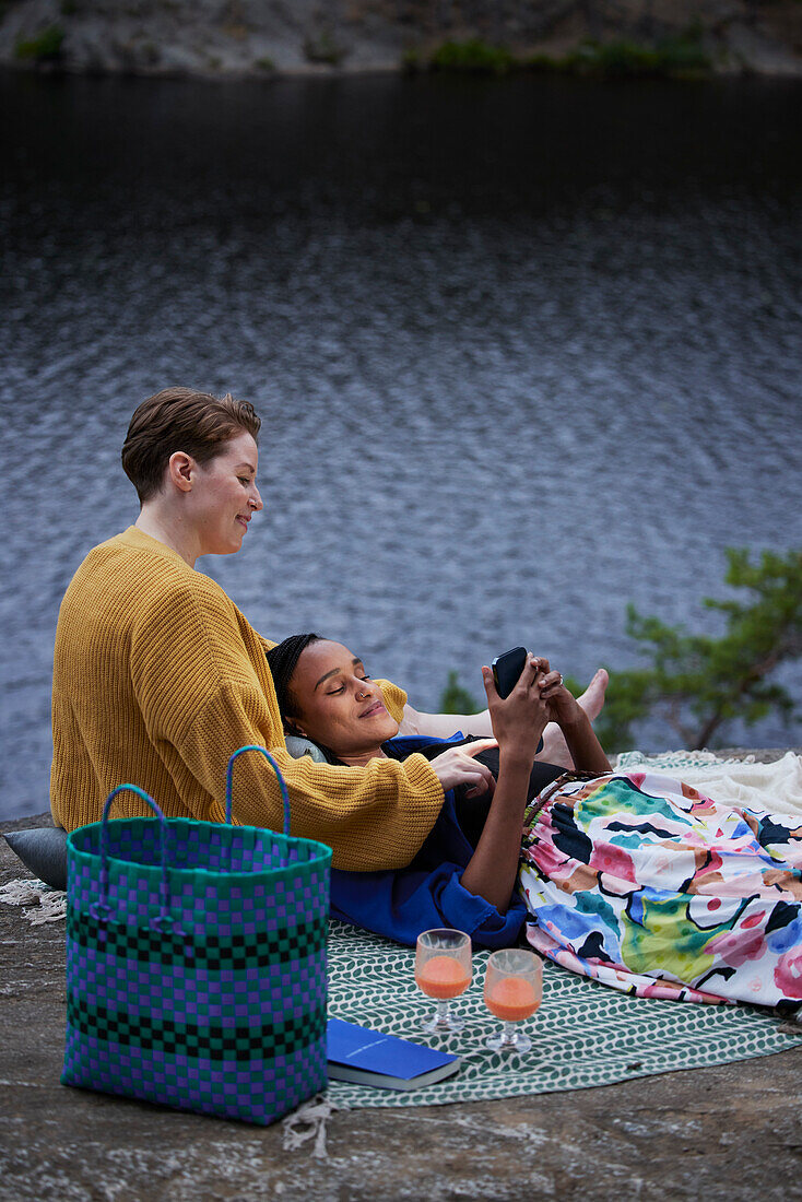 Female couple having picnic by river and using phone