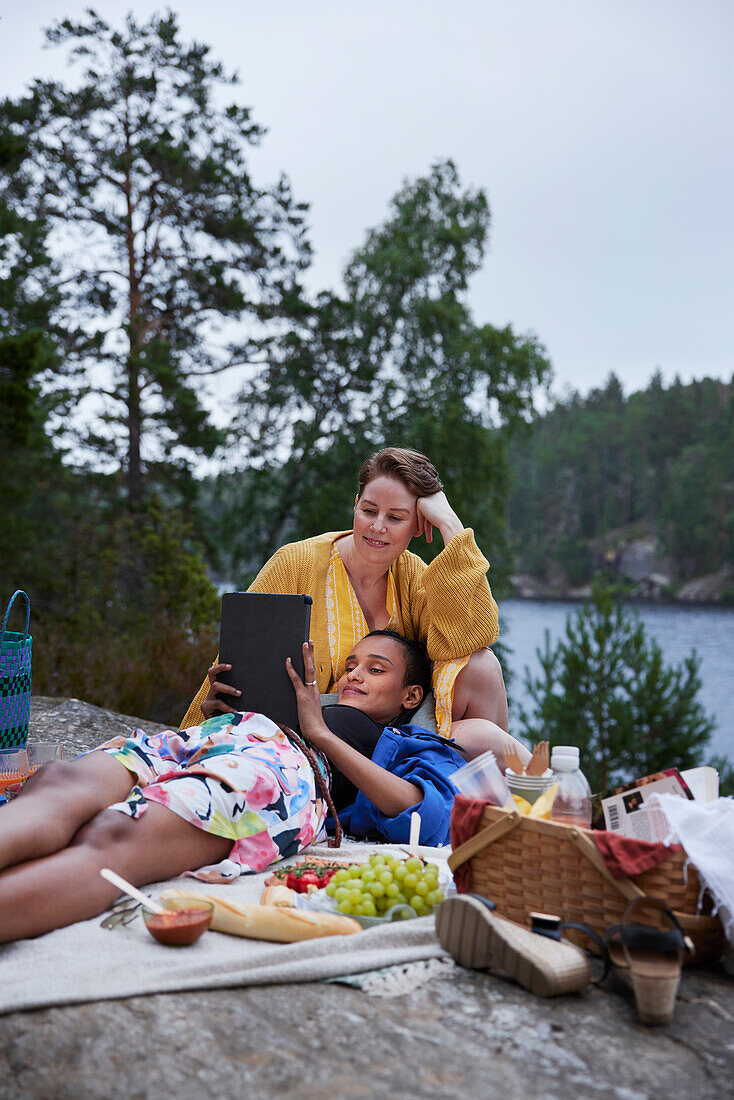 Female couple having picnic by river and using tablet