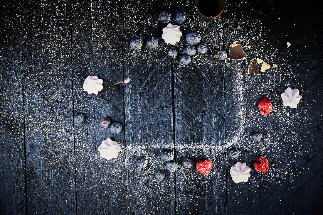 Puderzuckerrand, Baisertupfen und Beeren auf schwarzem Holzuntergrund