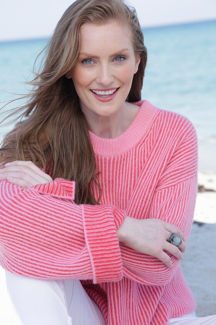 Young, blond woman in pink jumper and white trousers by the sea