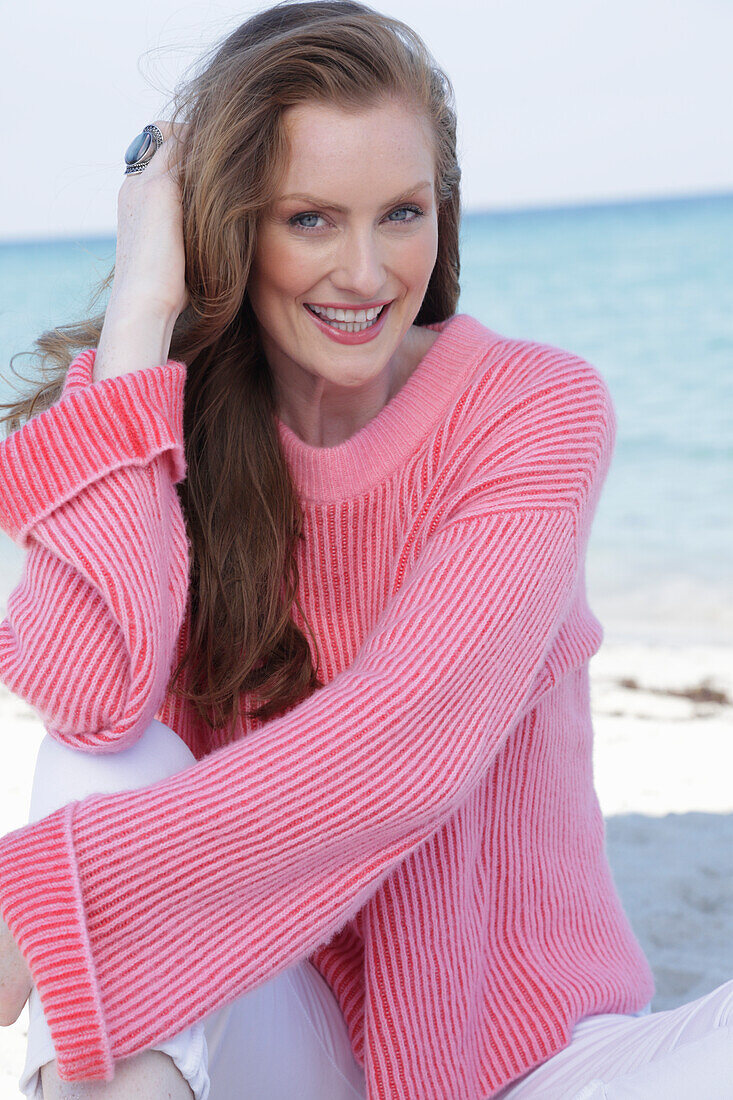 Young, blond woman in pink jumper and white trousers by the sea