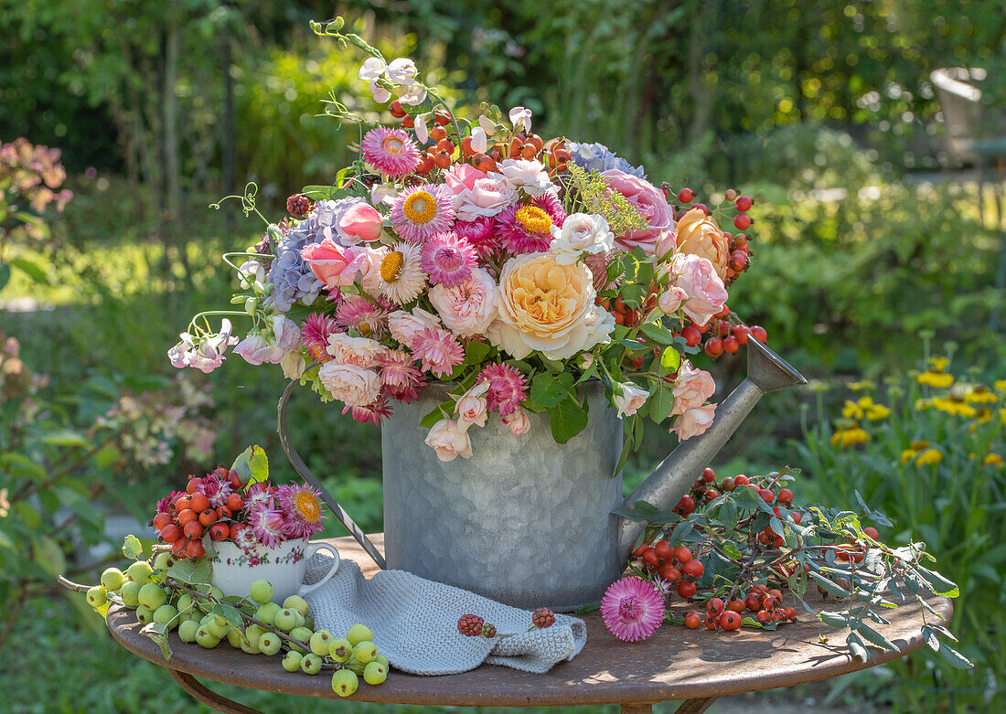Bouquet with roses, strawflowers, sweet peas, hydrangeas and rose hips in watering can on garden table