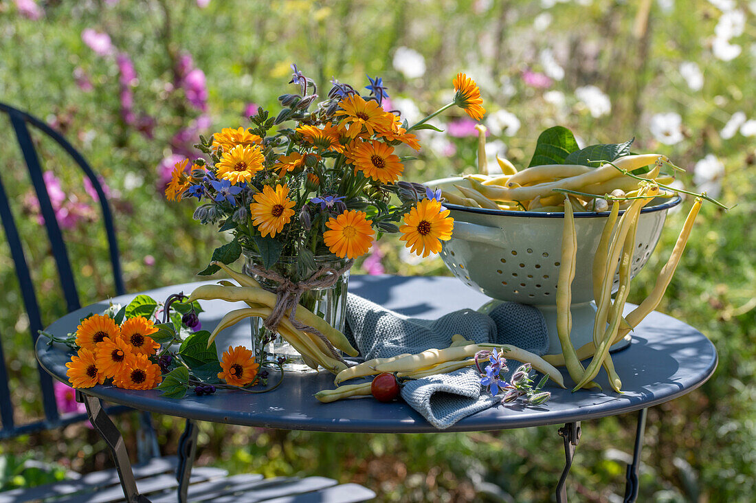 Ringelblumenstrauß mit Borretschblüten daneben Fußseiher mit frisch geernteten gelben Bohnen auf Tisch