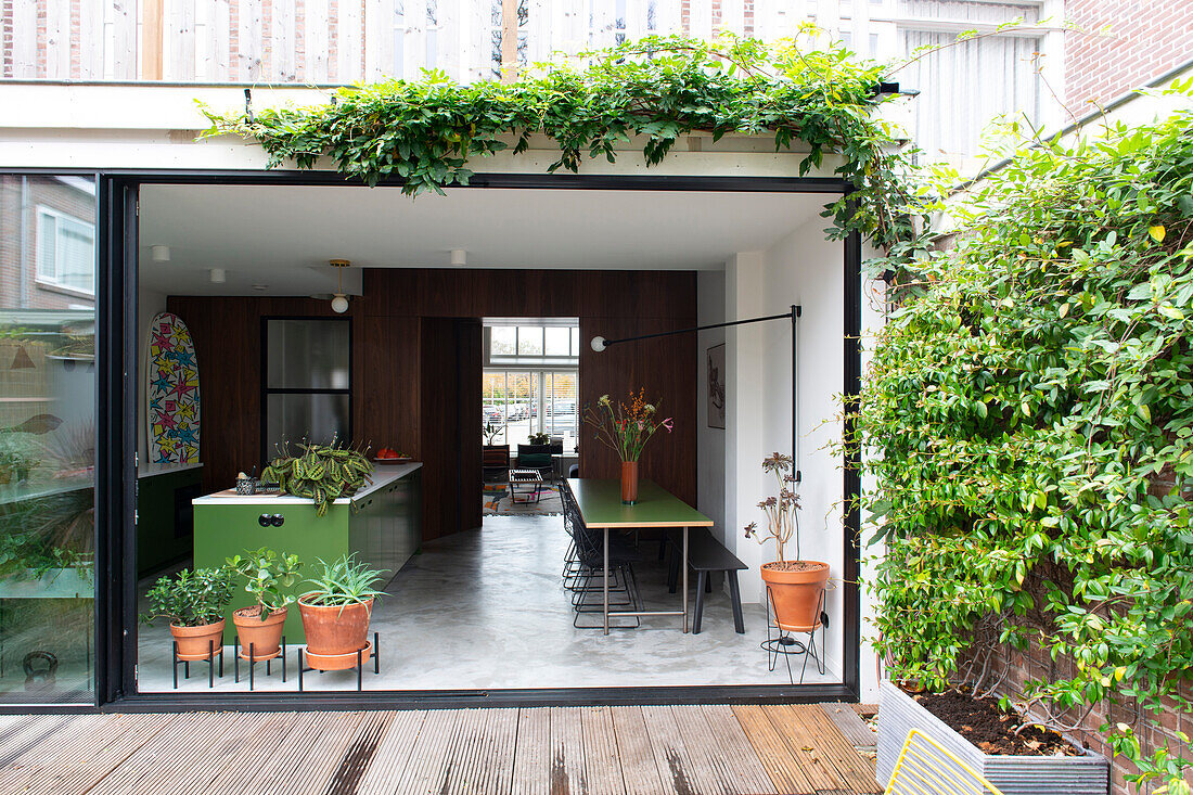 View through open patio door to a modern living room with kitchen and table in green and plants