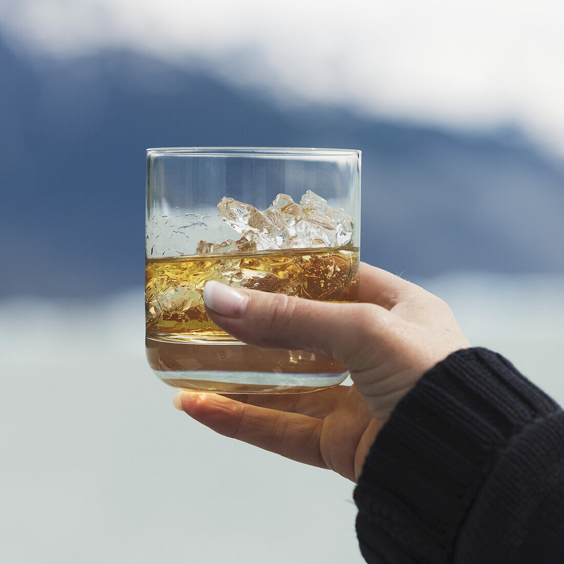 Die Hand einer Frau hält ein Glas mit Eis und einem Getränk, im Hintergrund eine See- und Berglandschaft; Provinz Santa Cruz, Argentinien