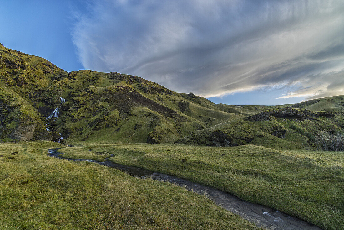 Linsenförmige Wolken bilden sich über einem Wasserfall und einem Bach, der in Richtung Meer fließt, in der Nähe von Vik; Island