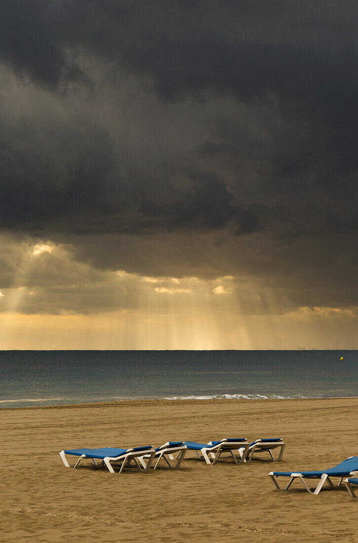 Sonnenstrahlen scheinen durch die dunklen Wolken über dem Ozean und Liegestühle am Strand; Benidorm, Spanien