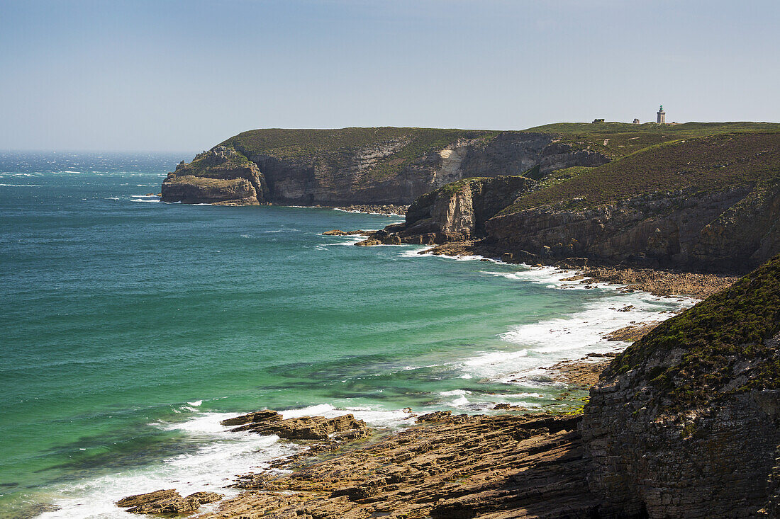 Felsige Küstenlinie und Bucht mit Turm im Hintergrund und blauem Himmel; Frehel, Bretagne, Frankreich