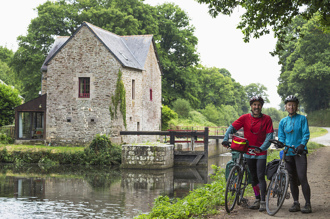 Couple With Bikes On A Path Along A Canal With Lock And Stone Lock House In The Background; Gaec Grand Boutron, Calorguen, Brittany, France