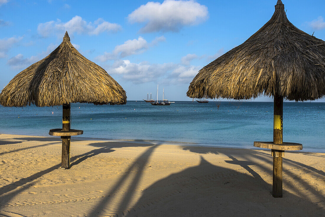 Strandpalapas am Palmenstrand mit Segelbooten, die nahe am Ufer verankert sind; Aruba