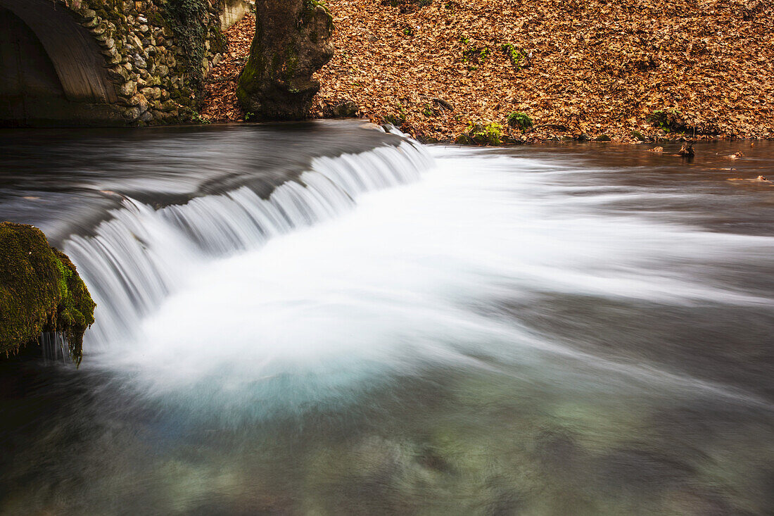 Water Cascading Over A Ledge In A River With The Riverbank Covered With Autumn Coloured Fallen Leaves; Naoussa, Greece