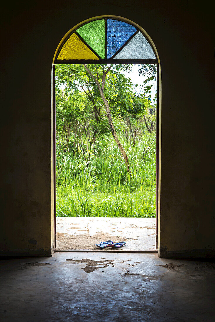 Ein Paar Sandalen an der Tür einer orthodoxen Kirche mit einem Buntglasfenster über dem Eingang; Gulu, Uganda